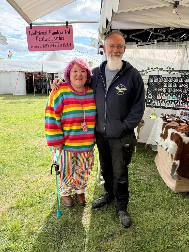 Dani (the Leather Lady) and Dave (The WiFi Guy), standing outside the DD Practical leather stall at a uk festival. It is gloriously sunny, the sky is blue and the grass is a lush green. Dani is wearing a stripy rainbow sweater and stripy rainbow trousers; she is leaning on her transparent blue walking stick. Dave has his arm around Dani and is wearing a dark blue hoodie and black combat trousers. They are both looking directly at the camera and smiling.