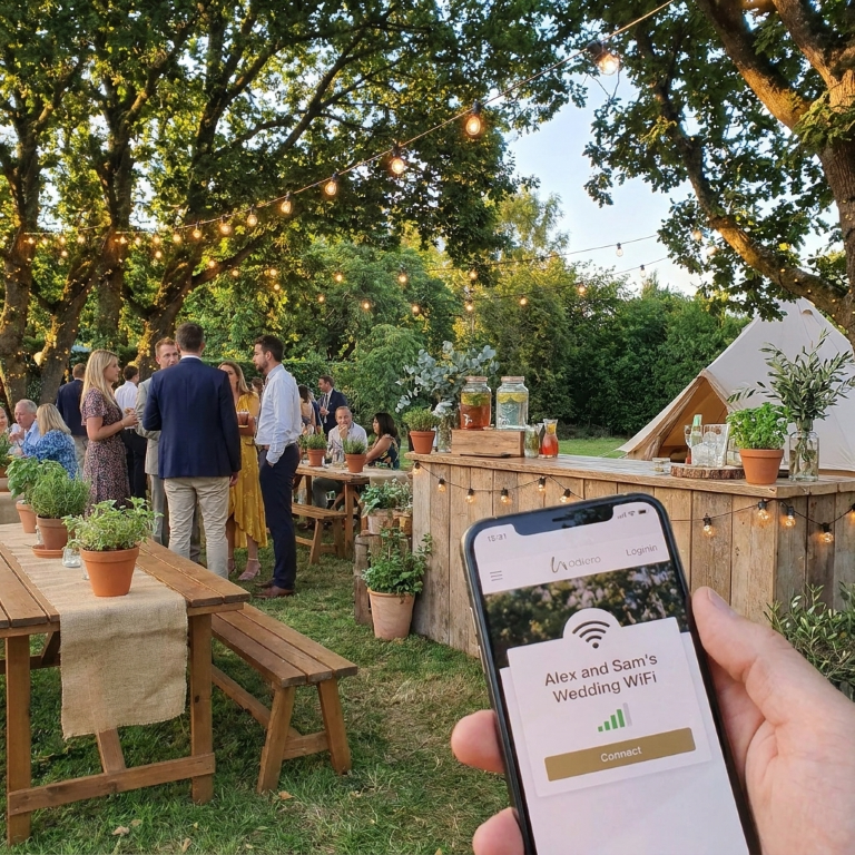 A mobile phone displays a customised WiFi captive portal at an outdoor wedding. The phone screen reads 'Alex and Sam's Wedding WiFi' with a Connect Button below it. In the background, guests mingle under festoon lights and trees in a lush, eco-friendly setting. A rustic wooden bar is visible in the background. Clear skys and golden hour lighting create a warm, welcoming atmosphere.