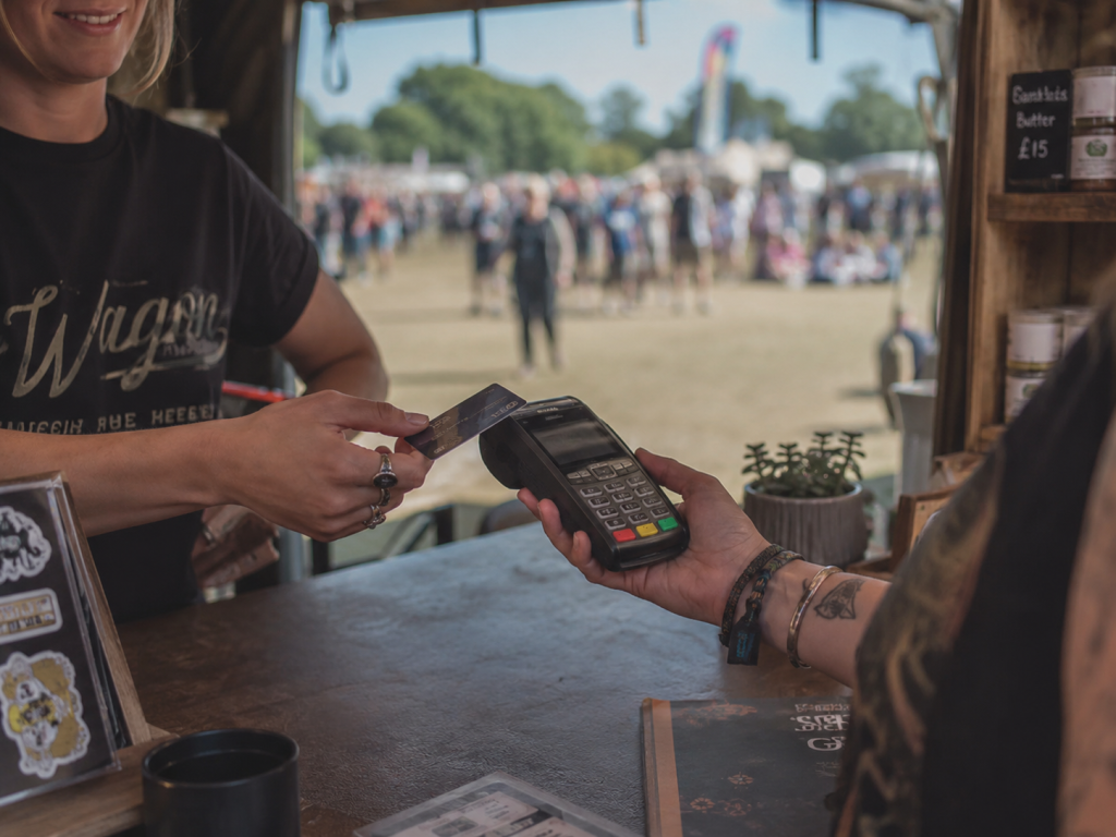 A Customer is making a card payment in a trade stall at a festival. The vendor is holding her card machine in her hand, we can see there is a small tatoo on her wrist and she is wearing several bracelets. The Customer is offering her card to the machine in order to complete a contactless transaction. Through the open door in the background, we can see crowds enjoying themselves; there are indistinct tents, stalls and flags in the distance.