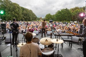 A Photograph taken from the back of the stage as Chesney Hawkes performs live at Lytham Hall on Saturday 26th August 2026. The back of the drummer is visible directly in front of the camera; the bass player is to the Right, and the guitarist is to the left. Chesney is Centre stage, holding his hand in the air and making a peace sign with his fingers. In the background, the audience can be seen dancing and returning the singer's gesture. Lytham Park stretches out into the far distance.
