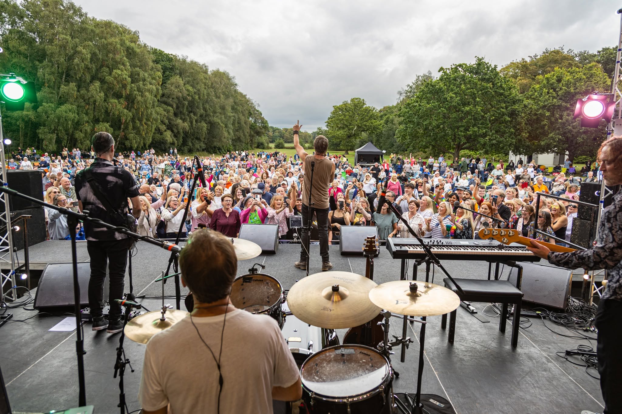 A Photograph taken from the back of the stage as Chesney Hawkes performs live at Lytham Hall on Saturday 26th August 2026. The back of the drummer is visible directly in front of the camera; the bass player is to the Right, and the guitarist is to the left. Chesney is Centre stage, holding his hand in the air and making a peace sign with his fingers. In the background, the audience can be seen dancing and returning the singer's gesture. Lytham Park stretches out into the far distance.